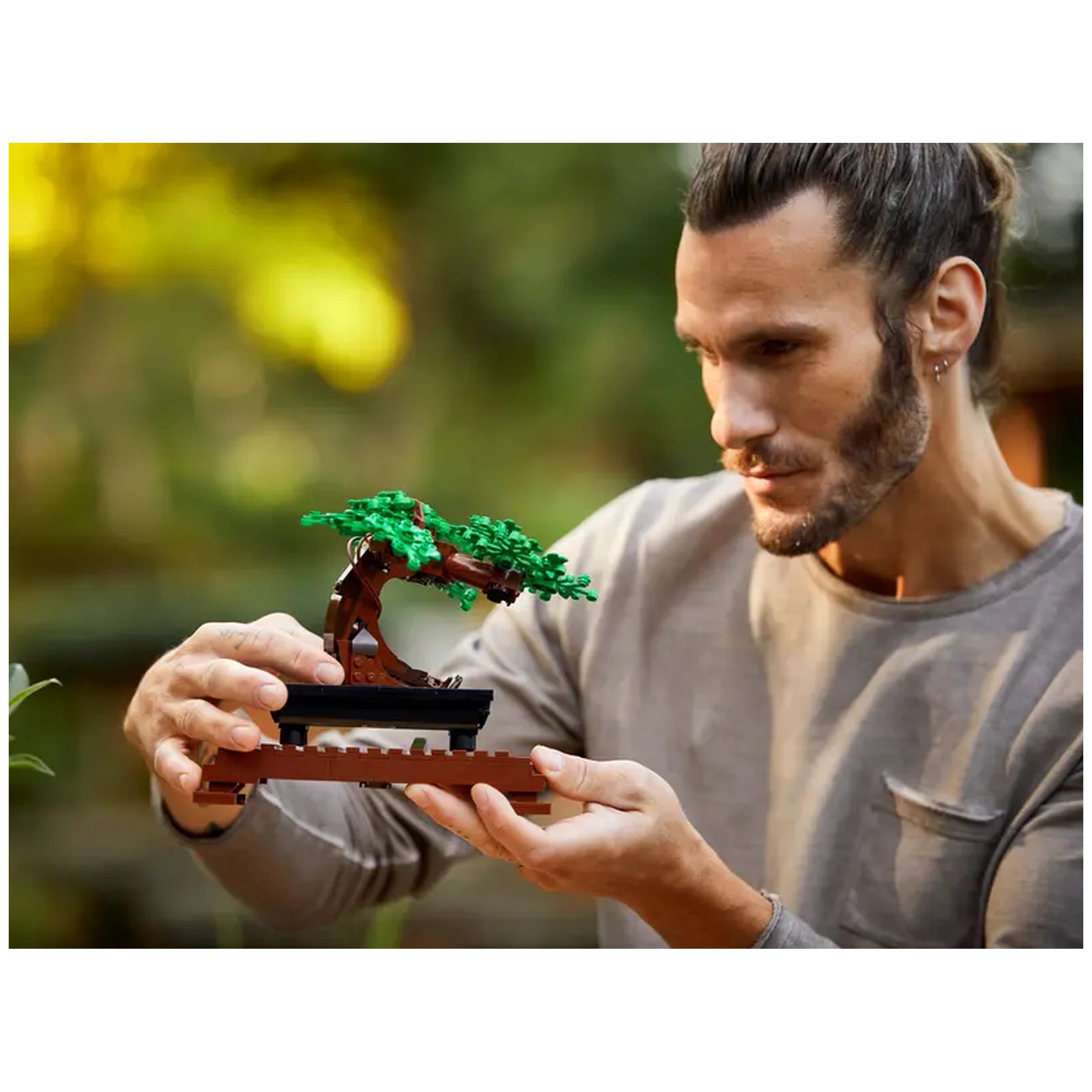 A man in a gray shirt delicately holds a colorful LEGO® Bonsai Tree model, showcasing its green leaves and intricately designed trunk, surrounded by a softly blurred green background.