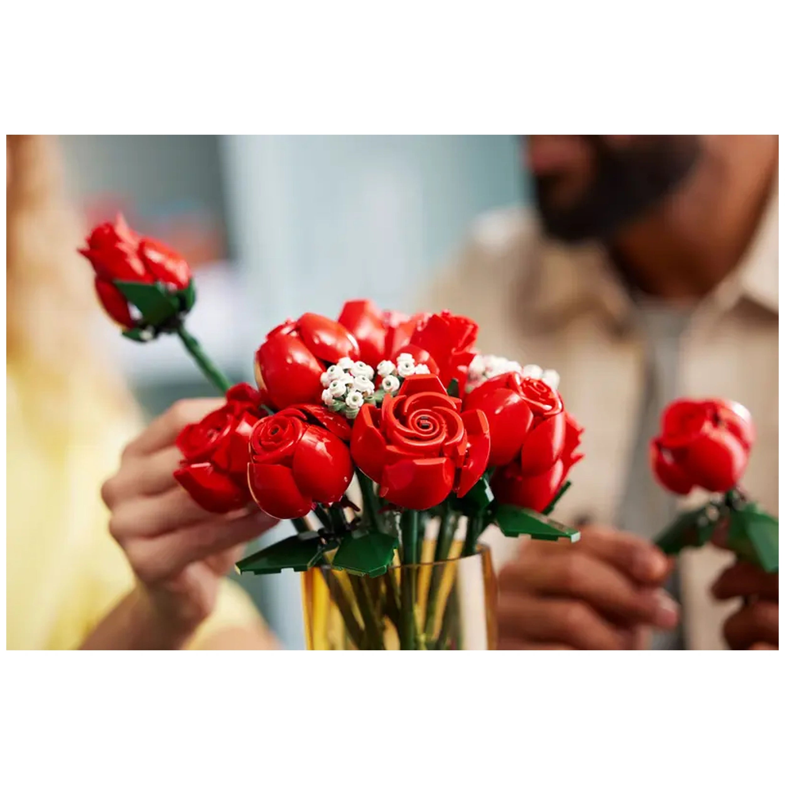 A vibrant, buildable bouquet of red LEGO® roses, featuring a mix of fully bloomed, blossoming, and budding flowers, accompanied by delicate baby's breath, held by a person in a light-colored shirt, with a blurred figure in the background.