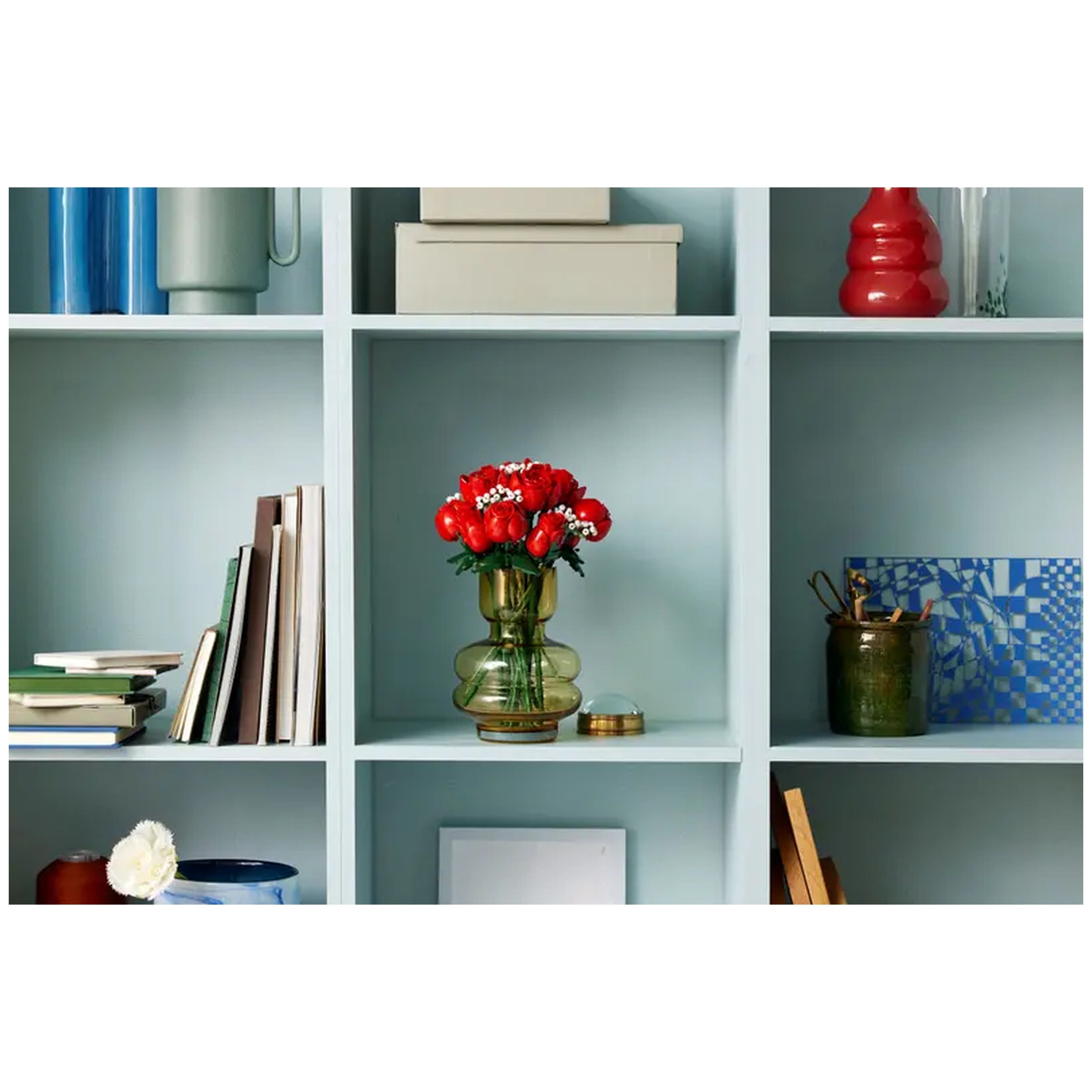 A vibrant bouquet of twelve red roses with small white flowers sits in a green glass vase on a light blue shelf, surrounded by books and decorative items.