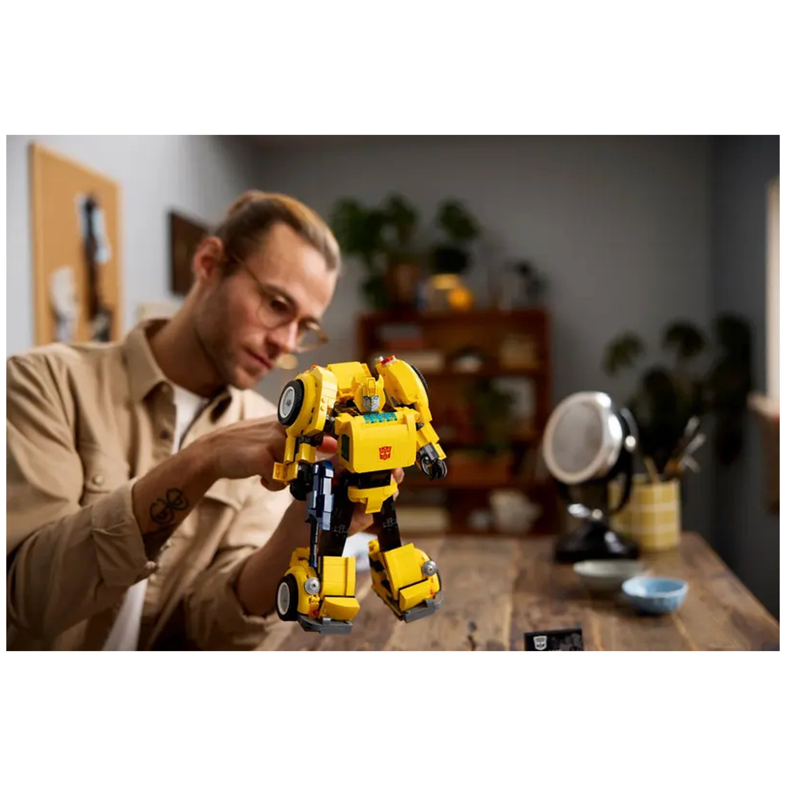 Adult man in a beige shirt carefully poses a detailed yellow LEGO Bumblebee robot model with articulated joints, set on a wooden table surrounded by plants and decorative objects.