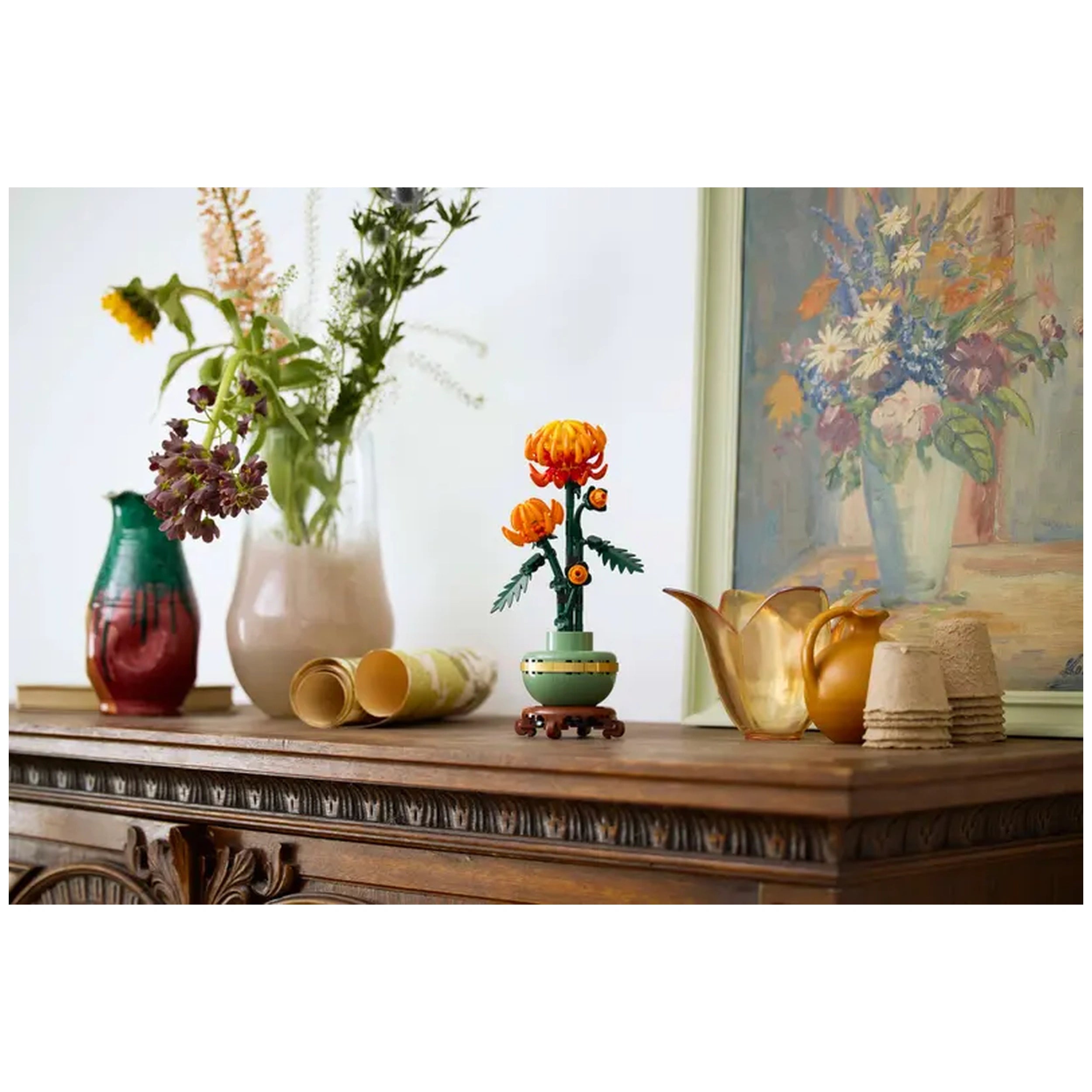 A vibrant LEGO® Icons Chrysanthemum set features orange flowers with posable petals in a green pot, displayed on a wooden table alongside colorful vases and rolled papers, creating a cozy home environment.