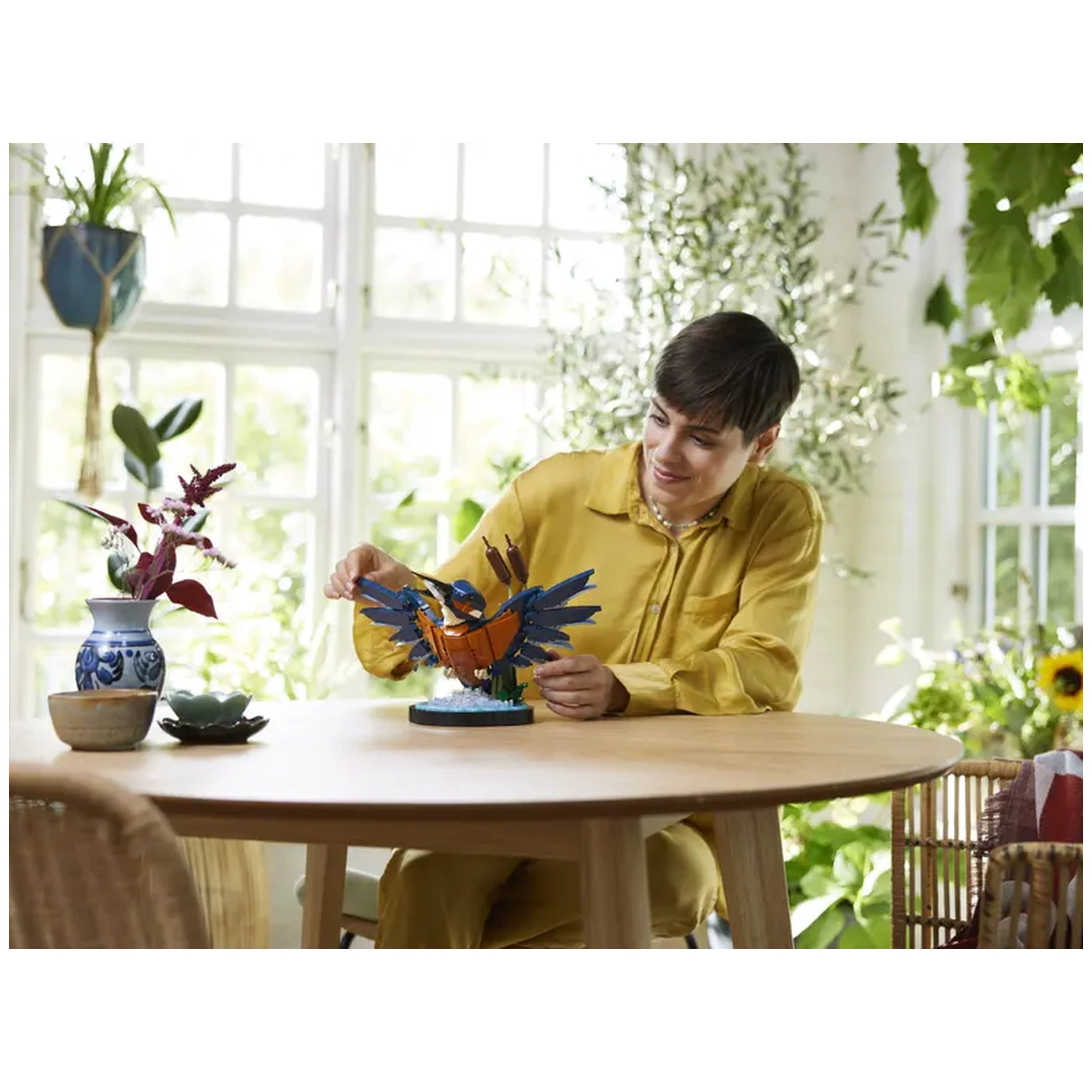 A person in a yellow shirt smiles while assembling the LEGO® Icons Kingfisher Bird set on a wooden table. The vibrant model features a kingfisher with blue and orange plumage, perched near buildable reeds and a water setting. Surrounding the scene are decorative plants and a vase with flowers.