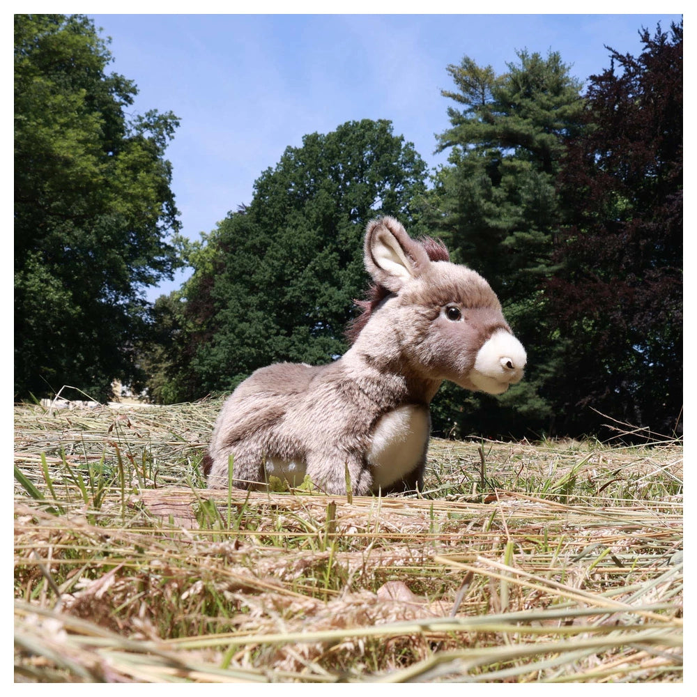 Eco-friendly plush donkey sits on straw in an outdoor setting with trees and a clear sky, showcasing a realistic texture.