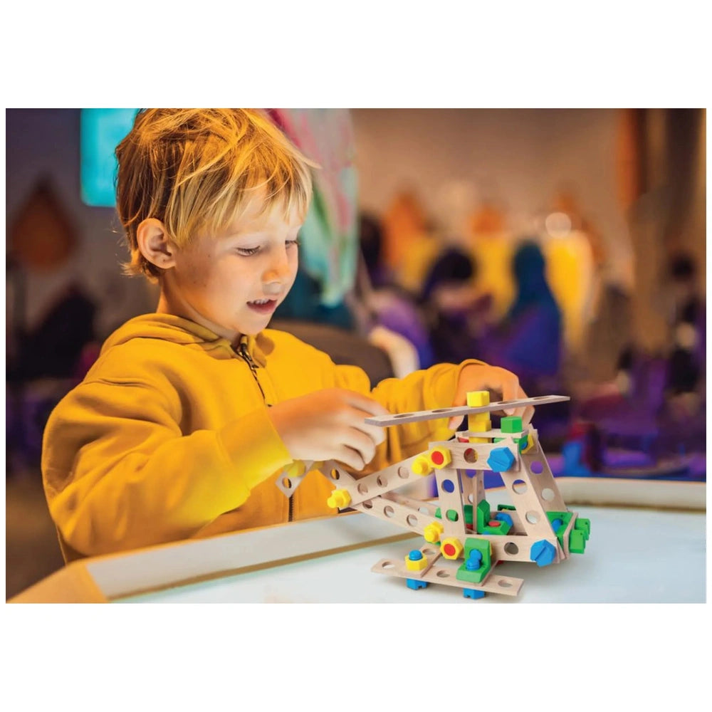 Young child in a yellow hoodie assembling the Alexander helicopter set with colorful screws and parts on a lighted table.