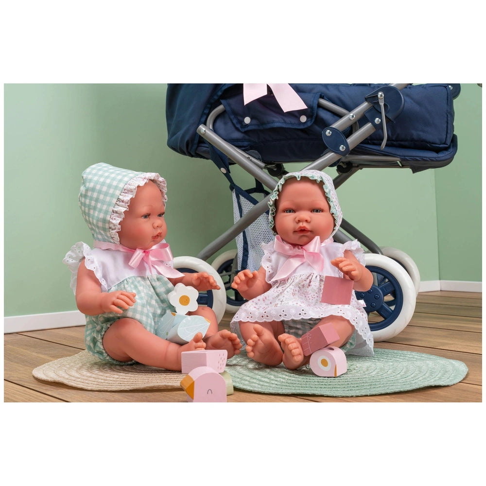 Two ASI Dolls in gingham and lace dresses sit on a rug with wooden blocks, beside a navy blue stroller.
