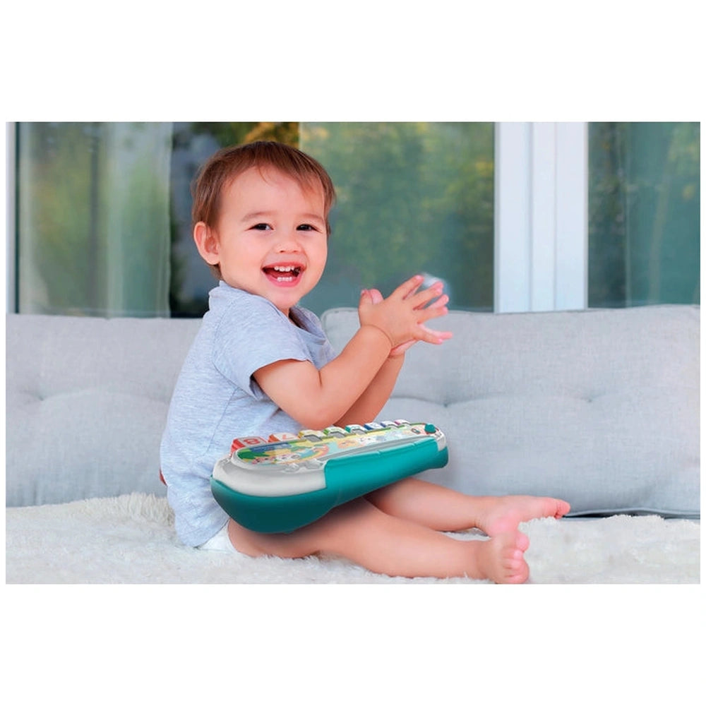Baby Clementoni Pianola Degli Animali toy held by a smiling child on a fluffy white surface, with greenery visible through a 