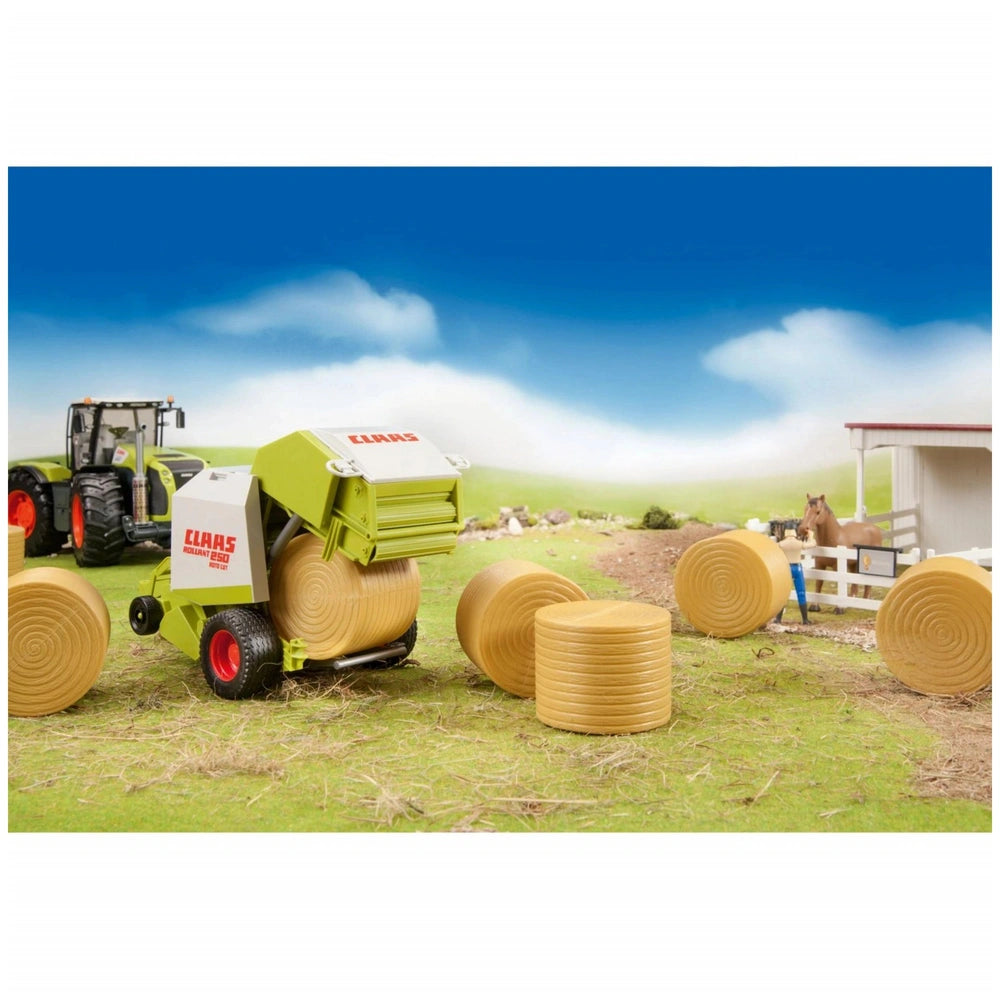 Toy Claas round baler on grass with hay bales, a tractor, and a horse stable in the background, under a blue sky.