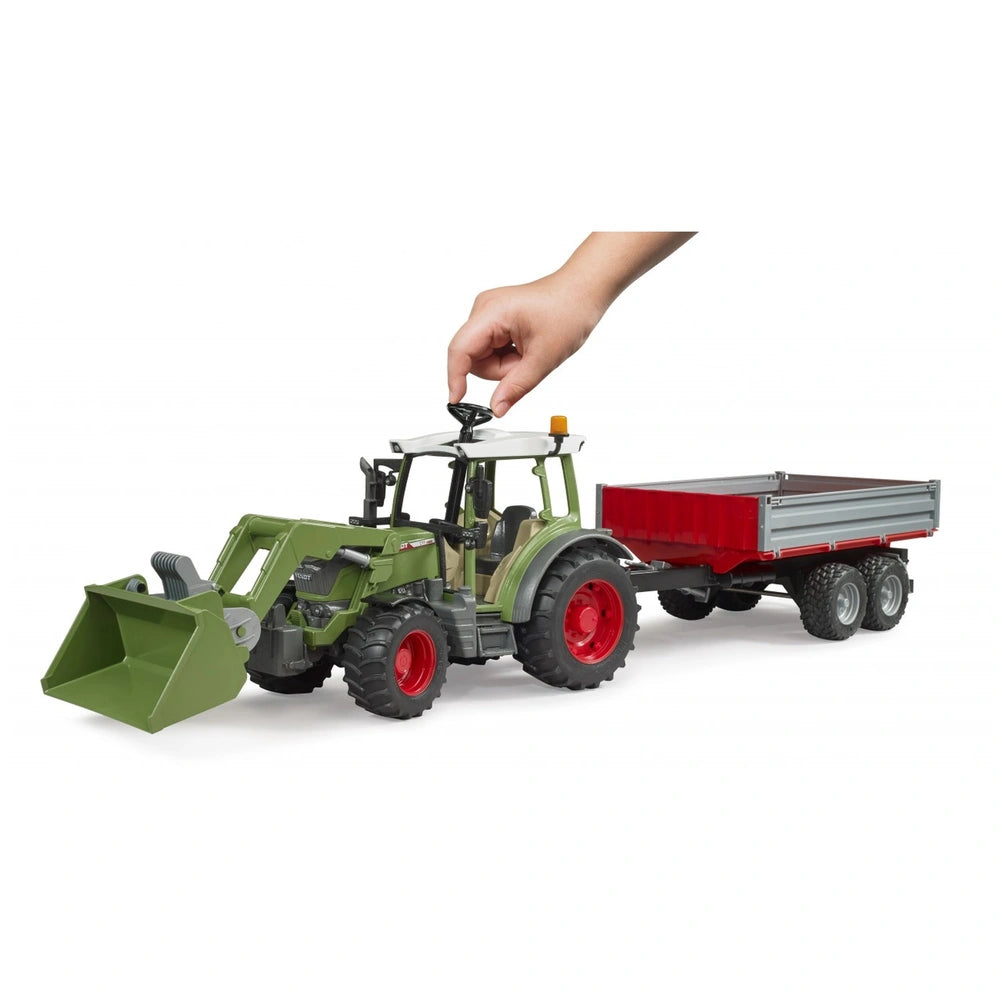 Green toy tractor with a frontloader and red tipping trailer, operated by a child's hand, on a white background.
