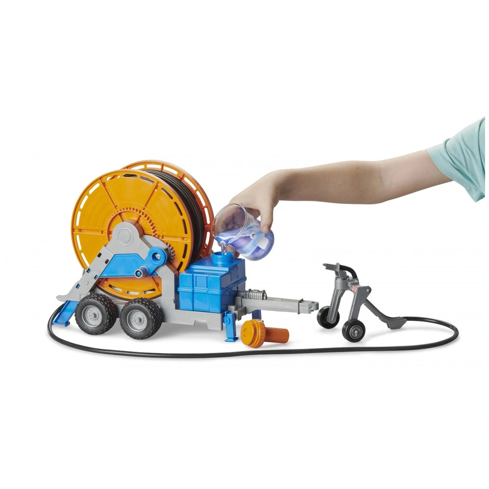 Child's hand pouring water into Bruder irrigation trailer toy with orange hose reel and blue tank.