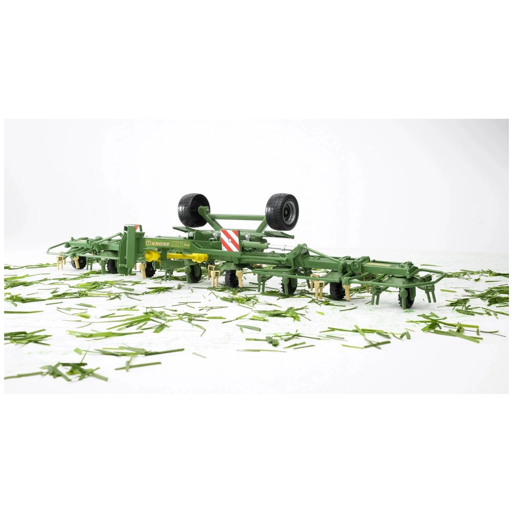 Bruder Krone Trailed Rotary Tedder toy, green with wheels, on white surface scattered with grass.