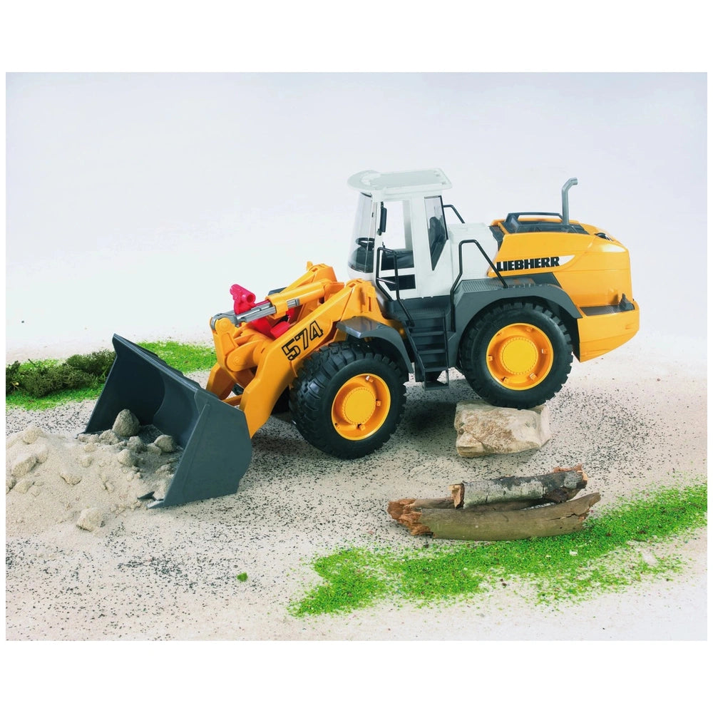 Bruder Liebherr L574 road loader toy with yellow body and tilting shovel, on sand with rocks and greenery.