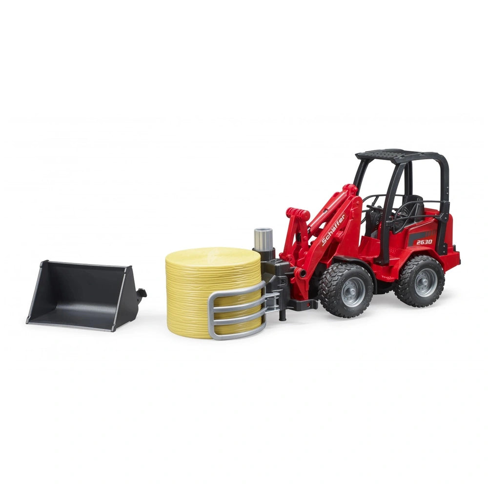 Red Bruder Schäffer Compact Loader 2630 toy with bale gripper holding yellow bale, detachable bucket nearby.