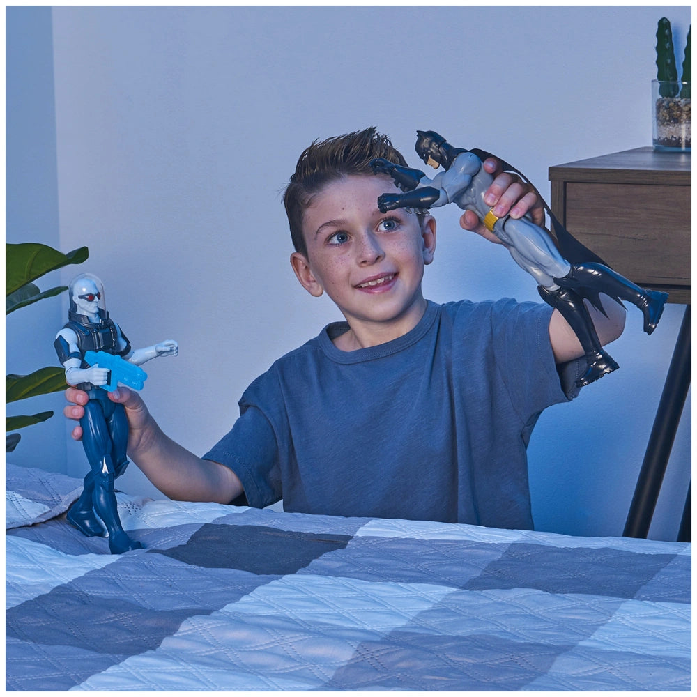 Batman Rebirth action figure held by a smiling boy in a blue t-shirt, with a checkered bedspread and plant nearby.
