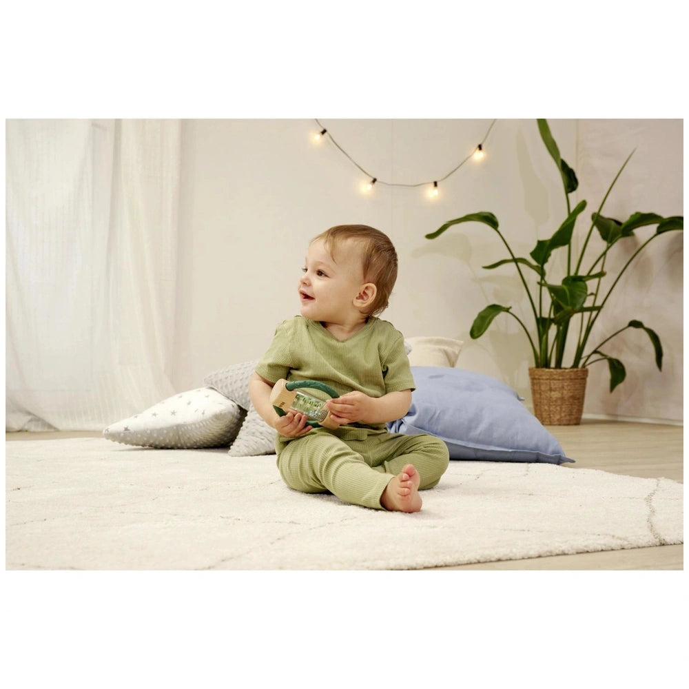 Eichhorn Baby HiPP Sound Rattle is held by a smiling child sitting on a plush white rug in a softly lit indoor setting.