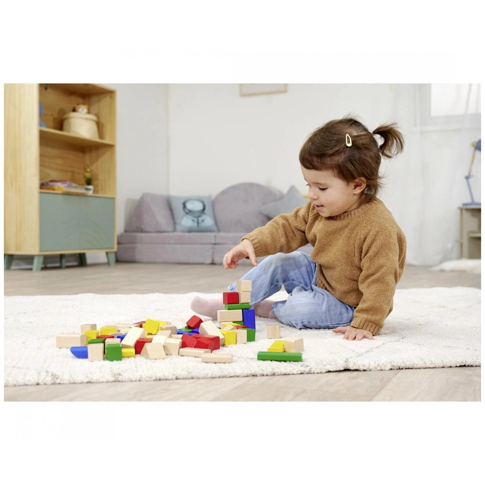 Eichhorn baby wooden coloured blocks scattered on a white rug, with a child in a brown sweater stacking them near a couch.