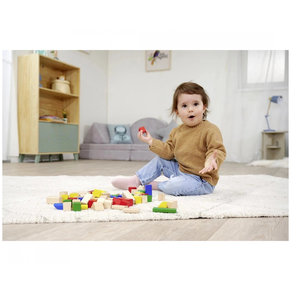 Eichhorn Baby Wooden Coloured Blocks scattered around a child on a white rug, with a gray sofa and wooden bookshelf in the ba