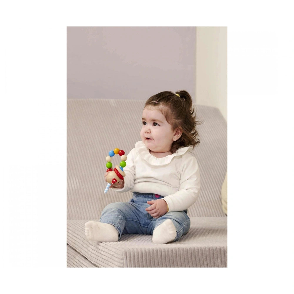 Baby wooden teething ring rabbit held by a child in a white top and blue jeans, sitting on a soft cushion.