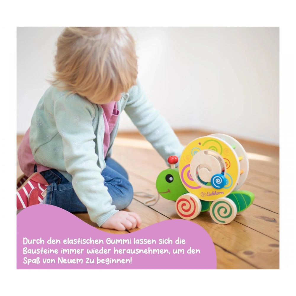 Colorful pull-along stacking caterpillar toy on a wooden floor, as a toddler in blue and pink plays nearby.