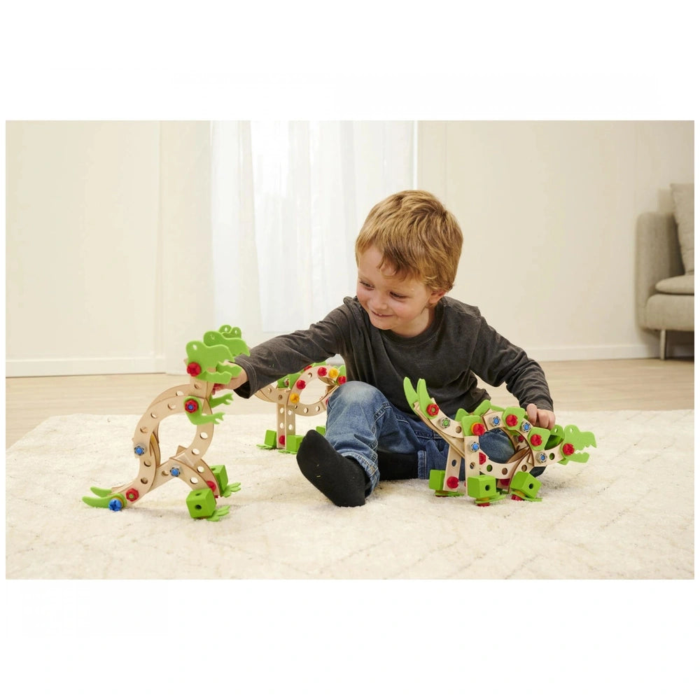A child with short blonde hair plays happily on a white rug with a green and brown wooden dinosaur construction set.
