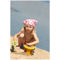 Gowi beach bucket in the foreground with a girl in a pink hat and red swimsuit playing on sand under a clear blue sky.