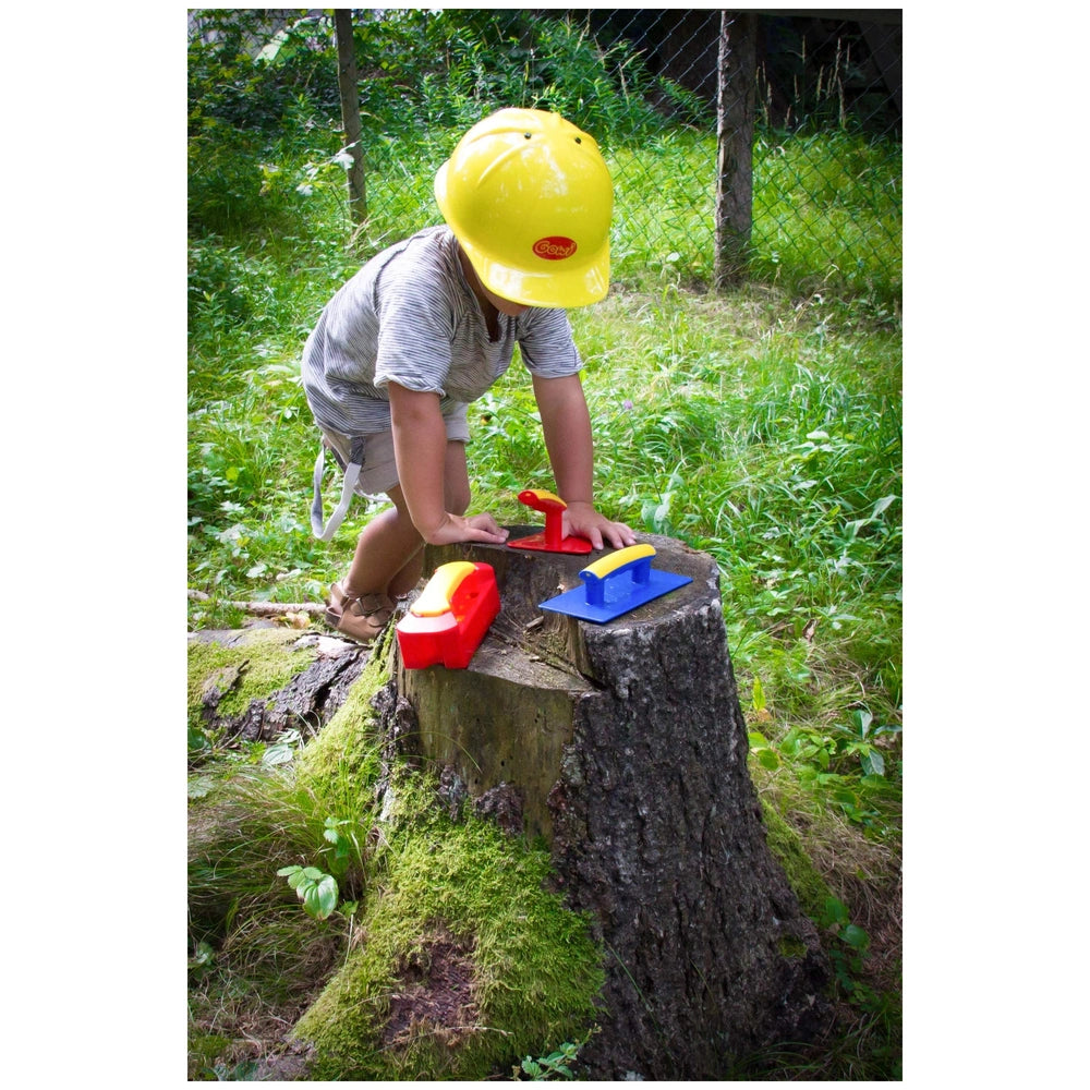 Gowi Design Bricklaying Set with a child in a yellow helmet, holding red and yellow and blue objects on a grassy stump.