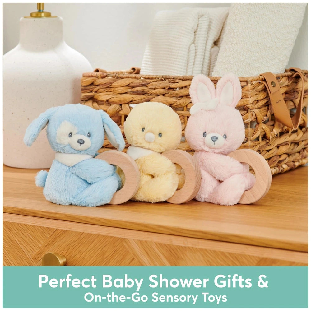 Gund Soft Animal Ring Rattle plush toys in blue, yellow, and pink arranged on a wooden surface with a wicker basket in the ba