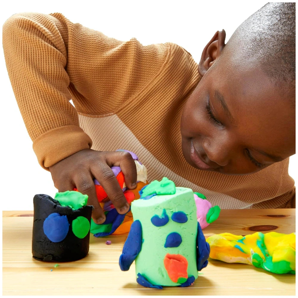 Child in a brown sweater joyfully plays with Hasbro Play-Doh 15 Cans Party Bag on a wooden table, fingers engaged with clay.
