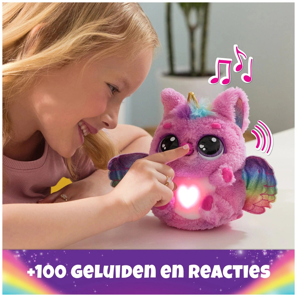 A young girl with blonde hair smiles at a pink plush Hatchimals toy with purple and blue details on a white surface.