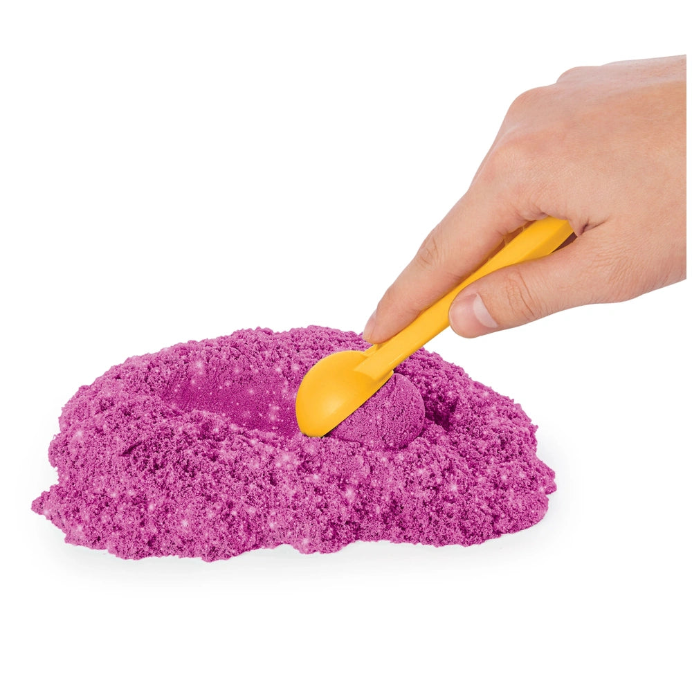 Kinetic Sand Shimmer Sparkle Sandcastle Set features a human hand holding a yellow spoon with purple sand on it.