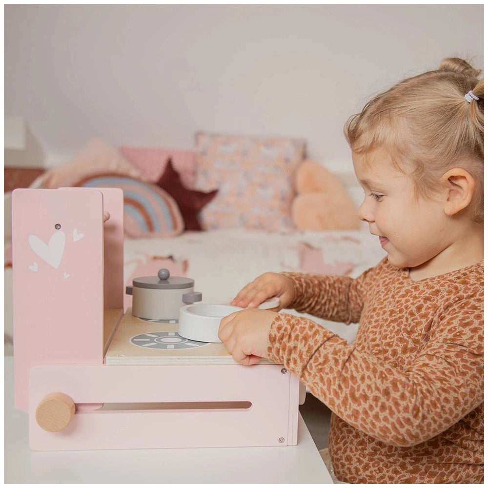 Child playing with a gray foldable cooker toy on a wooden table, surrounded by a bed of white pillows and stuffed animals.