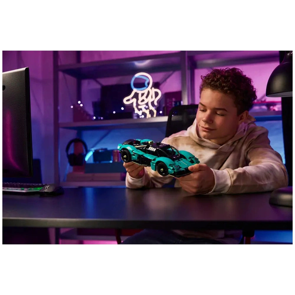 LEGO Aston Martin Valkyrie construction set toy held by a young male in a hoodie, sitting at a dark desk with ambient lightin