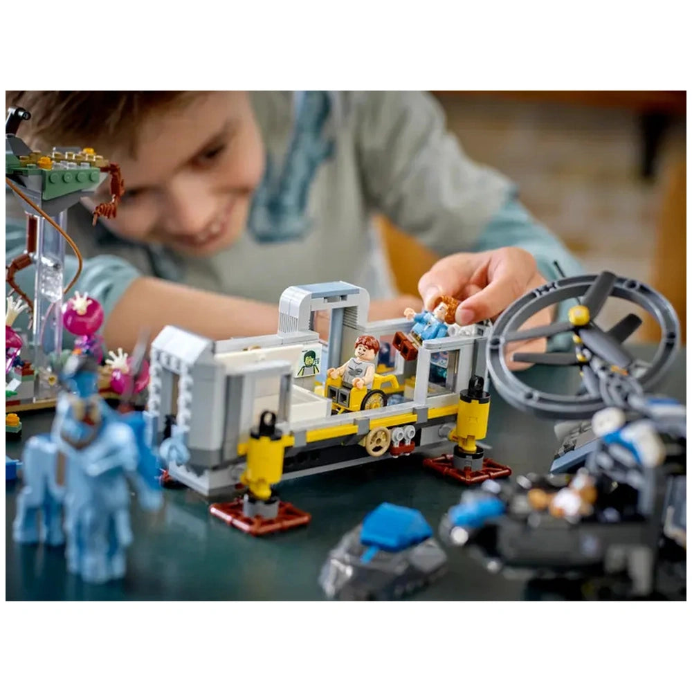 A child in a light blue shirt smiles while playing with a LEGO set on a dark table, surrounded by various LEGO pieces.