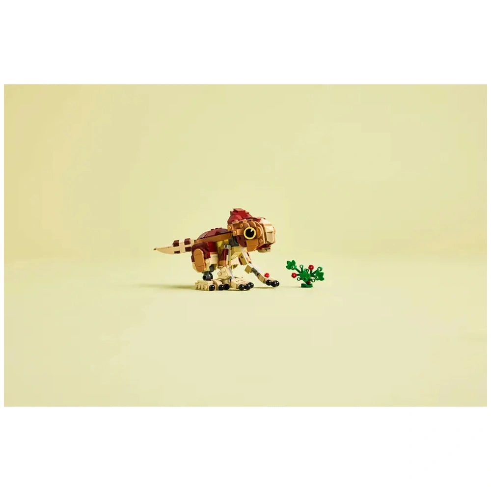 A colorful LEGO Baby Dinosaur Dolores figurine sits on a light background, playfully posed beside a small buildable plant with vibrant flowers. The dinosaur features movable parts, including arms, legs, and a tail, showcasing its intricate design.