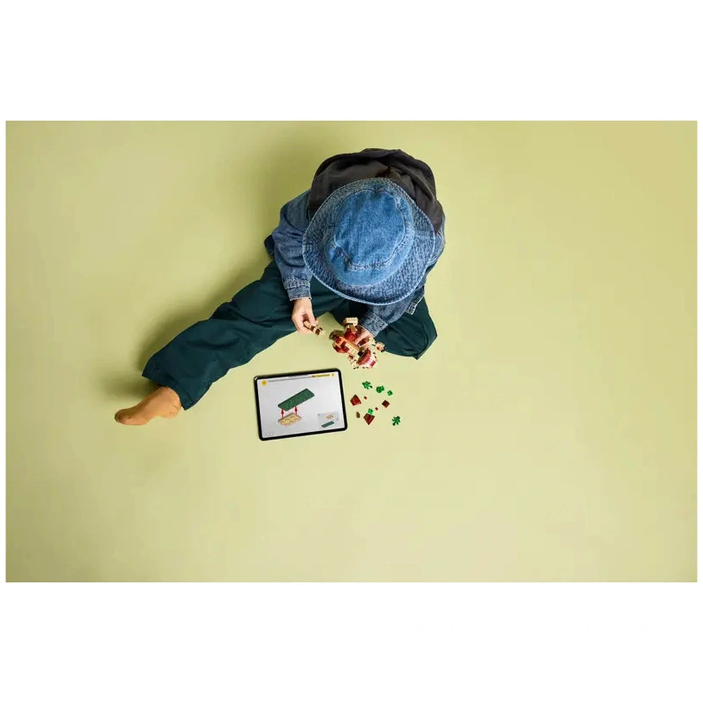 A young child sits on a light green floor, wearing a denim hat and jacket, assembling LEGO pieces. An interactive tablet displays building instructions, surrounded by colorful bricks.