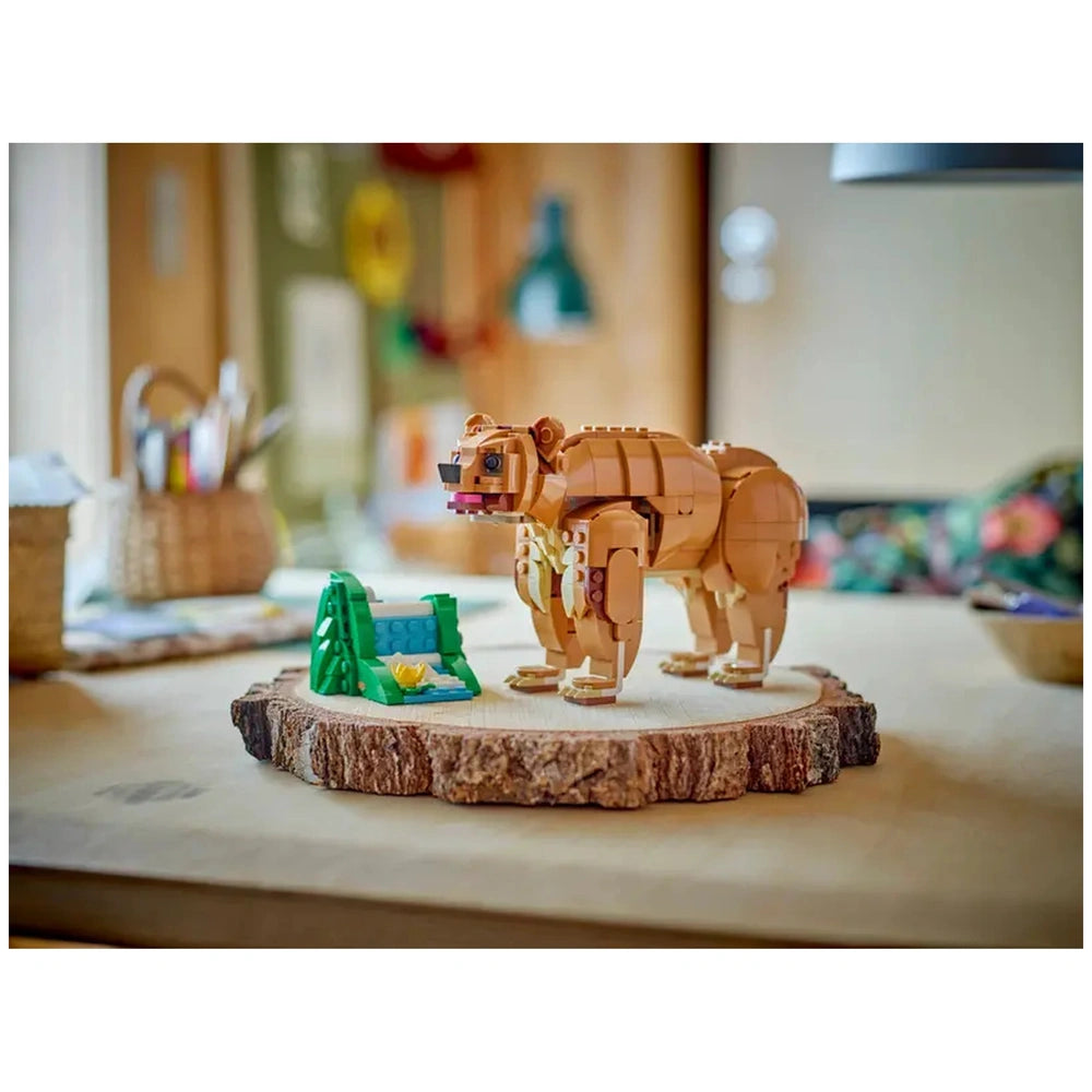 Posable LEGO bear figure in warm brown color, standing on a wooden slice base next to a green tent and yellow flower, with a cozy indoor background.