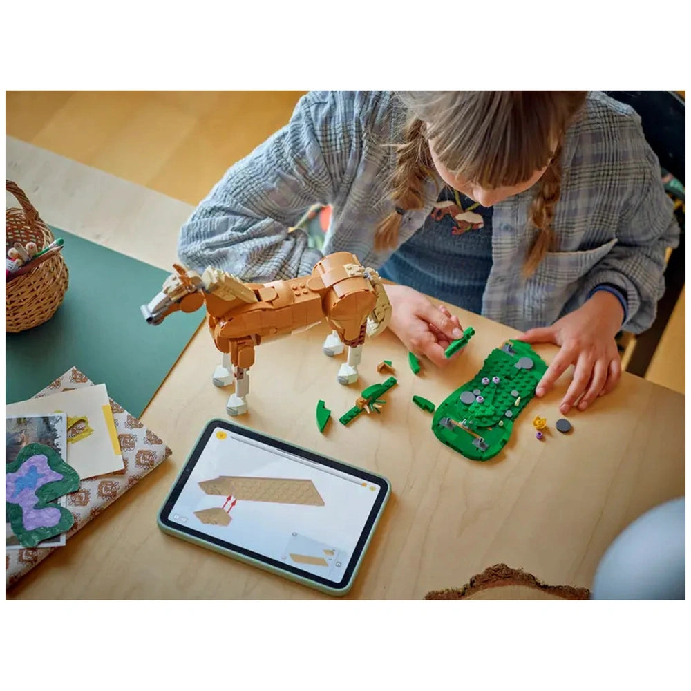 A child builds with LEGO pieces, arranging a brown horse figure and a green base with tree elements on a wooden table, while following instructions on a tablet.