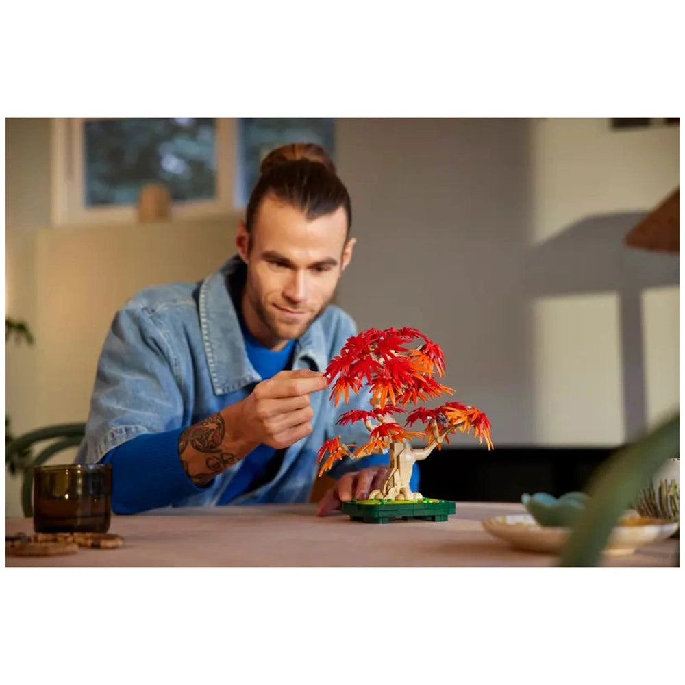 A man inspects the vibrant red and orange leaves of a LEGO Bonsai Tree on a tabletop, with a cozy interior in the background.