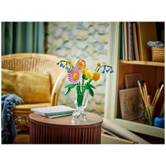 Colorful LEGO® Botanicals Petite Sunny Bouquet set displayed in a clear vase on a round wooden table, surrounded by a cozy living room.