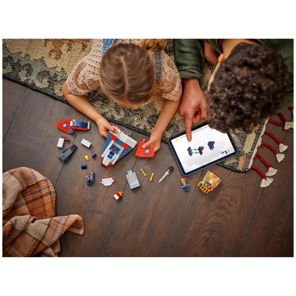 A child builds with LEGO pieces, holding a red and blue spaceship while an adult points to a tablet displaying construction instructions. Various colorful blocks and minifigures are scattered on a wooden floor beside them. A cozy blanket is draped nearby.