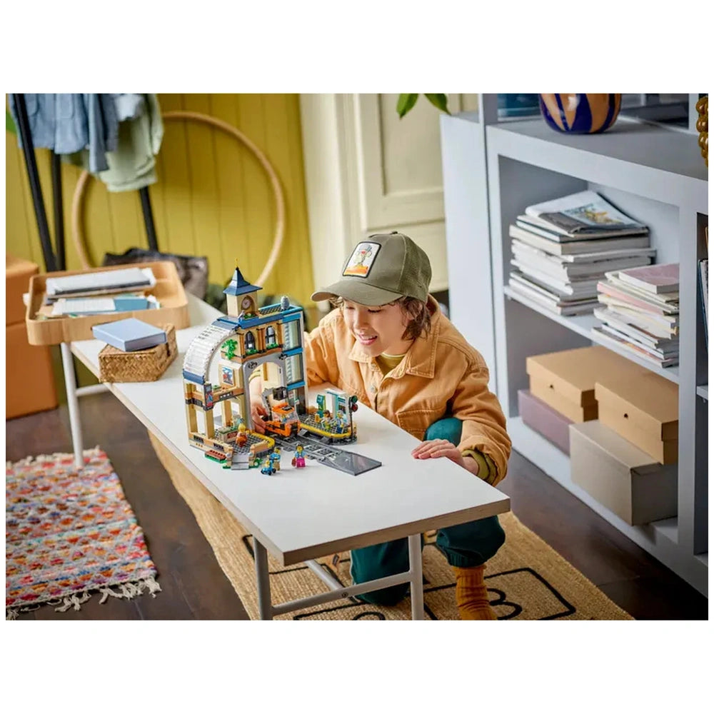 A child in a beige jacket and green cap interacts joyfully with the colorful LEGO Central Train Station set on a white table, surrounded by various miniature figures and accessories. The cozy room features a bookshelf filled with books and a desktop organizer nearby, adding a vibrant, playful atmosphere.