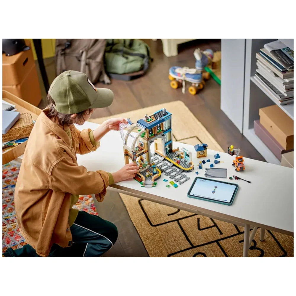 A child builds the LEGO City Central Train Station on a white table, surrounded by colorful blocks and a tablet, in a cozy room.