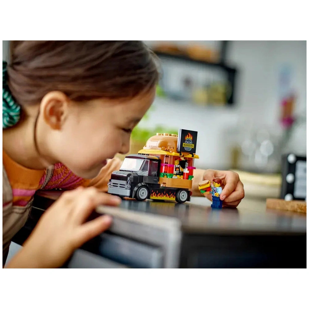 A young girl leans over a colorful LEGO® City Burger Truck, playfully serving a burger to a minifigure. The truck features a giant burger on top and vibrant food details.