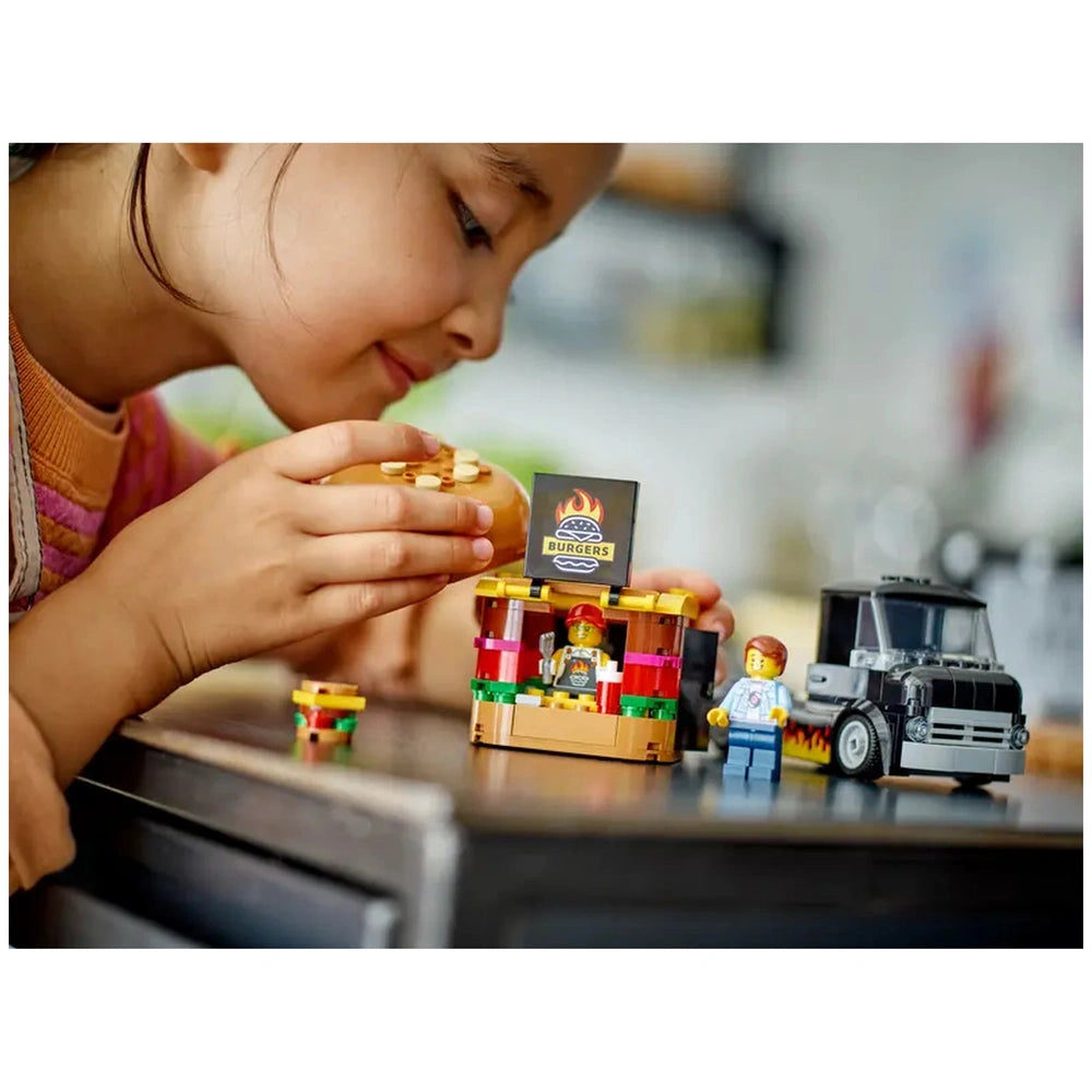 A child closely examines a LEGO® Burger Truck set, holding a miniature burger near a colorful food stand. The scene includes two minifigures: a vendor in front and a customer beside a black toy truck, with a vibrant "BURGERS" sign.
