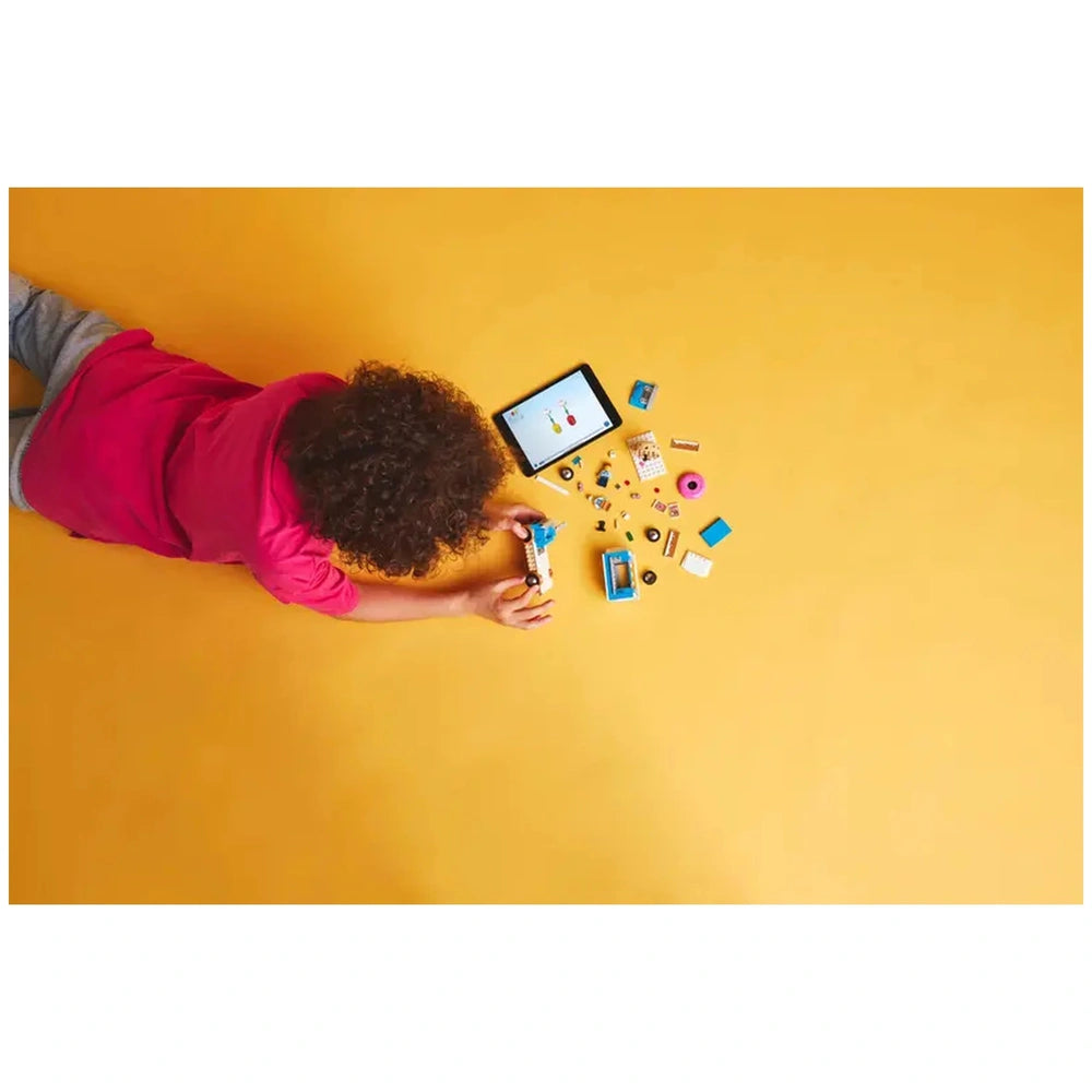 A child in a pink shirt lays on a yellow floor, focusing on colorful LEGO pieces and a tablet displaying building instructions.