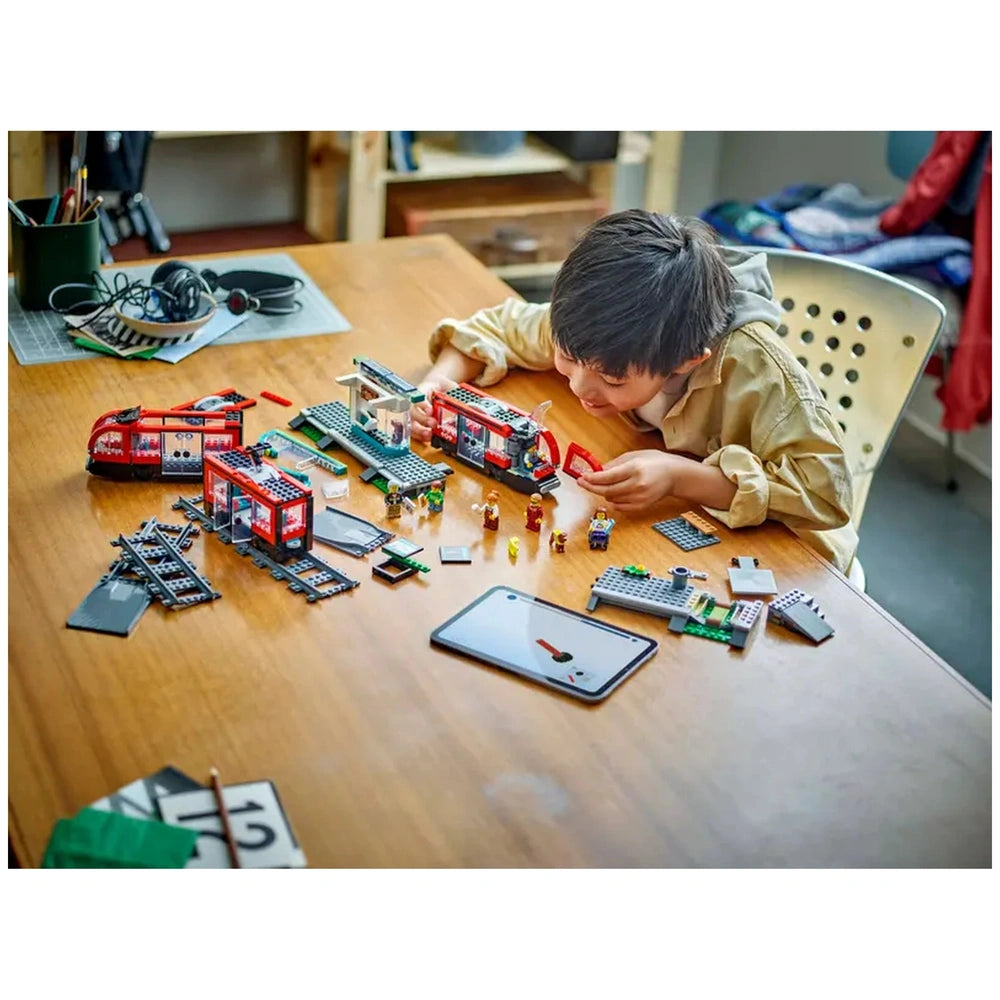 A young boy is assembling the LEGO® City Downtown Streetcar and Station set on a wooden table. Colorful streetcars, minifigures, and tracks are scattered around him, alongside a tablet and stationary items. He focuses intently on connecting pieces, showcasing creativity and engagement in imaginative play.