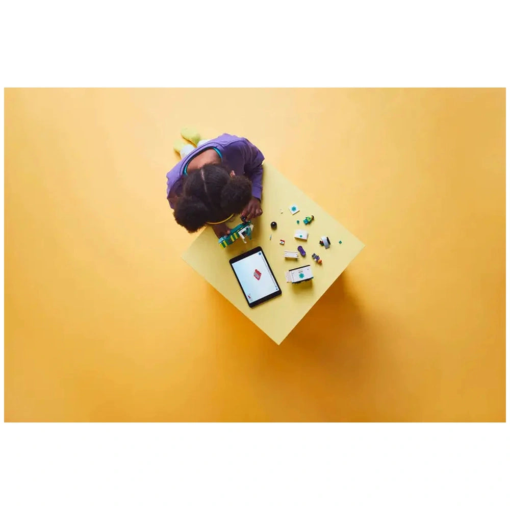 A child with curly hair focuses on building a LEGO ambulance at a yellow table, surrounded by colorful pieces and a tablet displaying instructions.