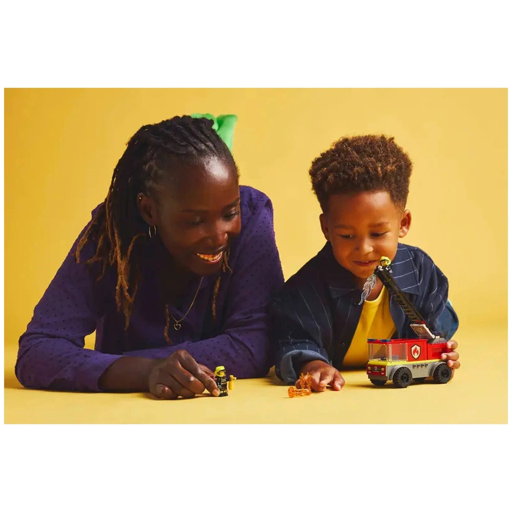 A smiling woman and a young boy play together on a yellow background, focusing on a LEGO City Fire Truck set and firefighter minifigures.
