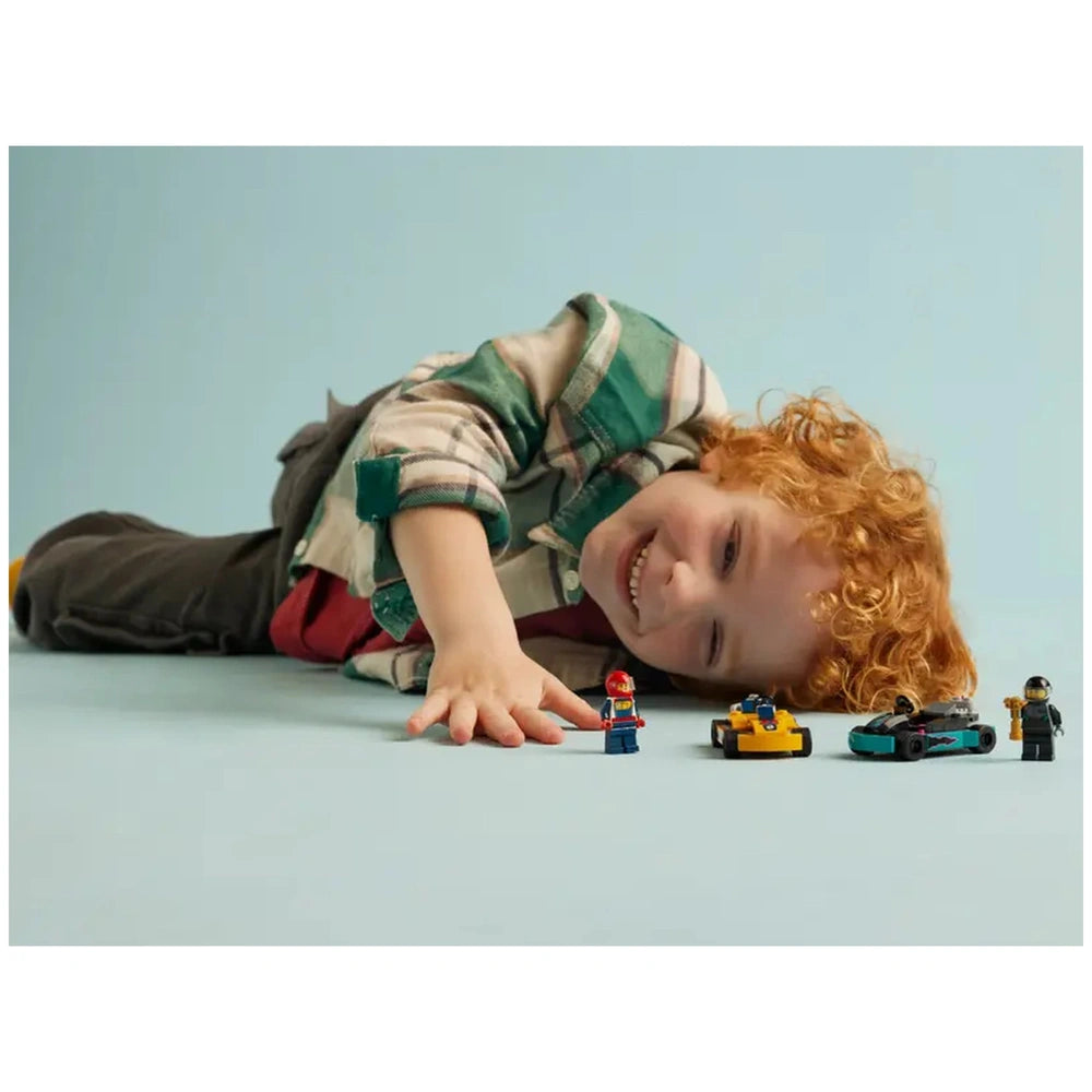 A joyful child with curly red hair lies on a light blue surface, reaching towards two mini LEGO go-karts—one orange and one turquoise—while holding a race driver minifigure. Another minifigure stands beside the go-karts, emphasizing the fun of the LEGO City Go-Karts and Race Drivers set.