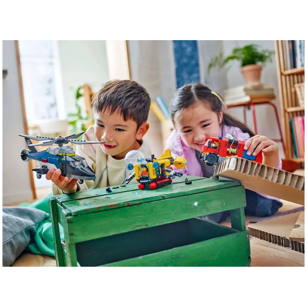 Two children play with LEGO® City toys on a green wooden table. One child holds a helicopter, while the other has a fire truck. A colorful submarine is also displayed on the table. The background features a cozy living space with natural light and bookshelves.