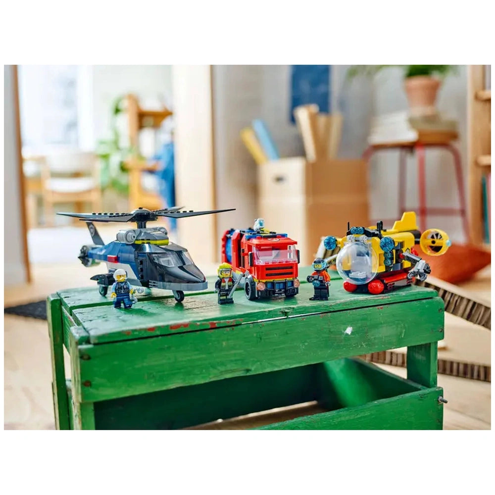 LEGO® City set featuring a police helicopter, fire truck, and exploration submarine on a green wooden crate, surrounded by LEGO® mini-figures.